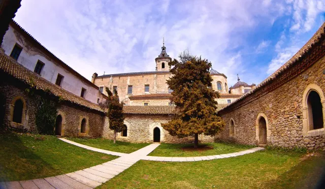 A stone monastery courtyard with arches, a paved path and a single tree on a green lawn