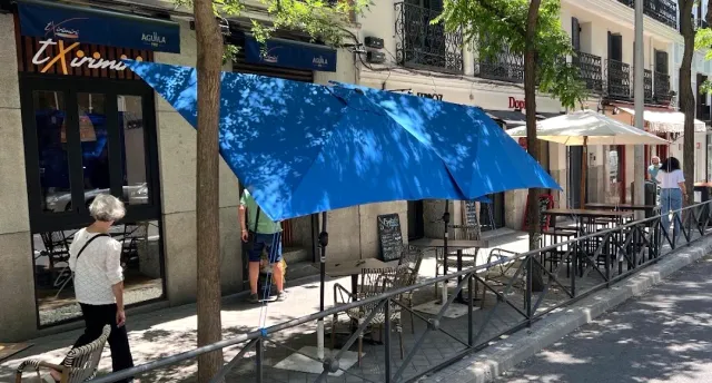 A pavement terrace with a blue parasol and metal tables, and passers by on a shaded street