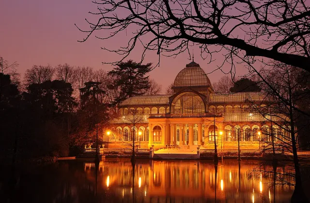 A glass pavilion with a dome lit from within at dusk, reflected in the pond before it