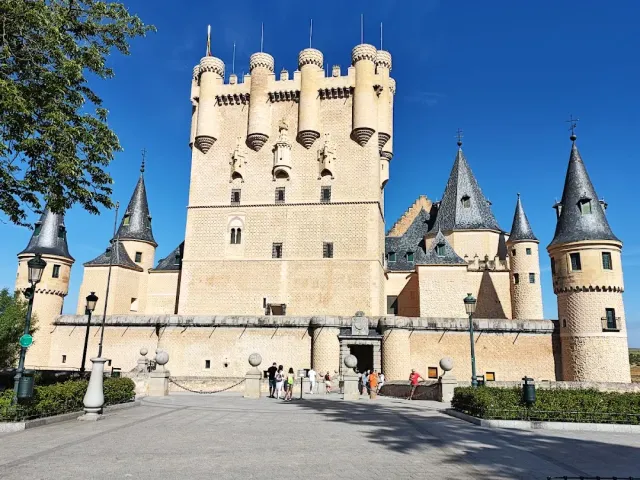 A pale stone fortress with dark pointed turrets against a blue sky, and visitors at the gate