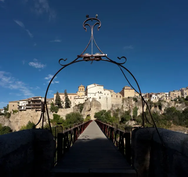 A narrow iron footbridge crossing a deep gorge towards a town built on a cliff, seen through an ornate iron arch