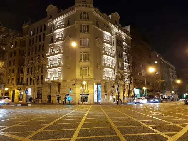 A lit corner building at night with balconies, facing a wide junction with yellow road markings
