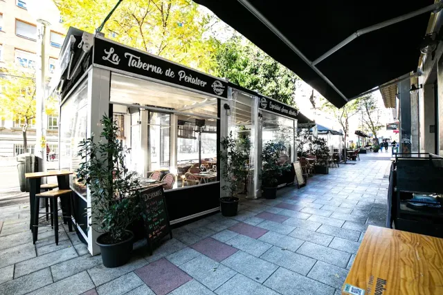 A glass enclosed terrace with a black awning, potted plants and a chalkboard on a wide pavement