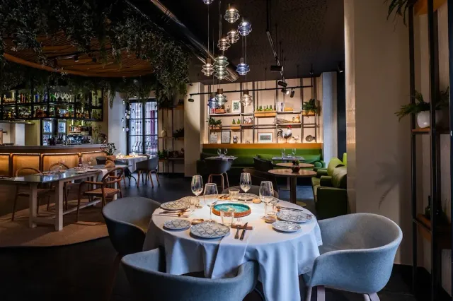 A restaurant interior with hanging greenery, glass lamps, a wooden bar and a laid round table