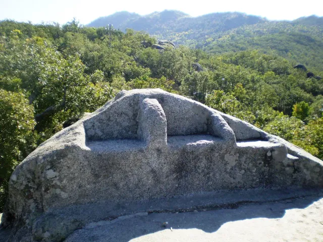 A large granite outcrop with stone seats carved into it, surrounded by trees and a mountain slope
