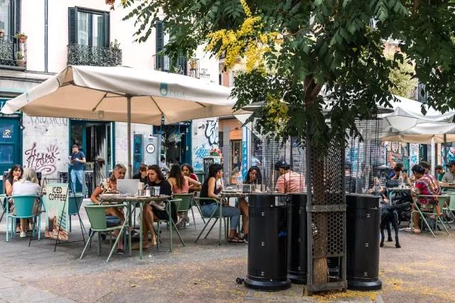 A street terrace with parasols and green chairs, and diners at tables under a tree
