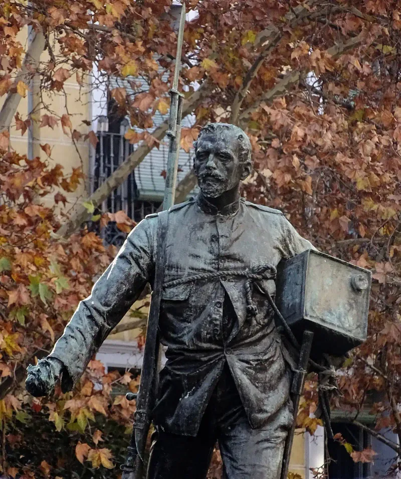 A bronze statue of a soldier holding a canister and a rifle against brown autumn leaves and a Madrid building