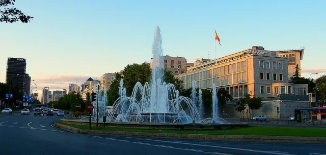 A large fountain with water jets in a round plaza, with buildings and trees around it