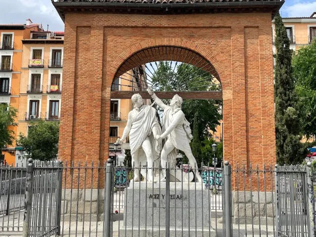 A brick arch with a marble statue of two figures at its centre, surrounded by an iron fence