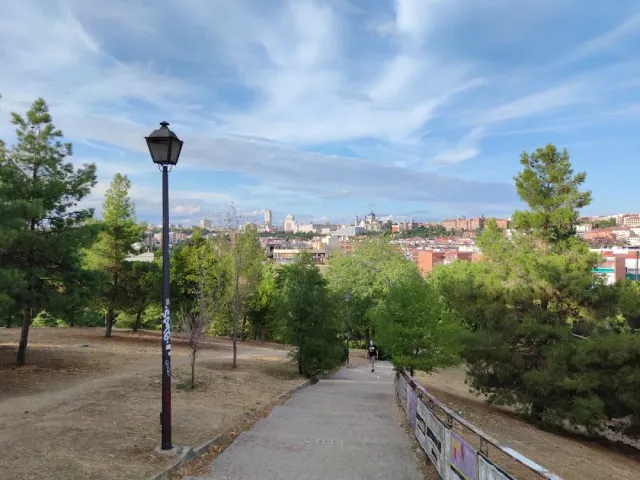 A park path with a tall lamp and pine trees, and the city skyline on the horizon
