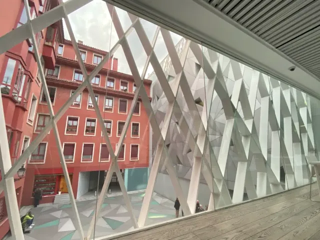 A view through a glass facade with a white lattice of bars toward a red building across the plaza