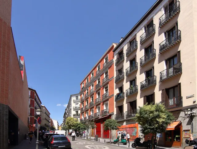 An uphill street with residential buildings, ground floor shops and trees on a sunny day