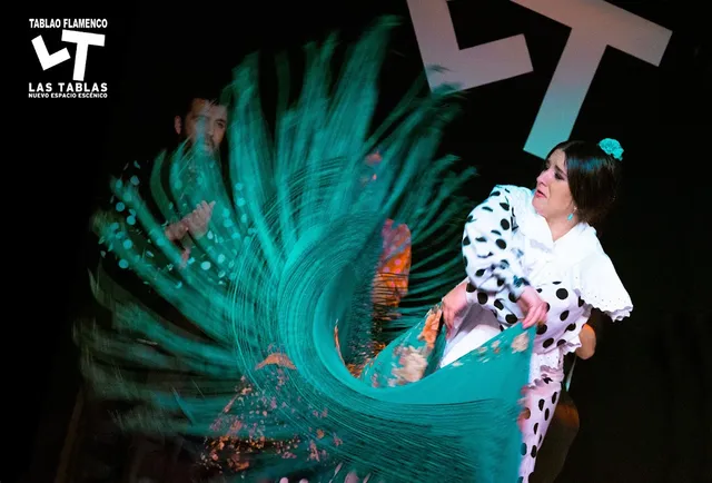 A flamenco dancer in a polka dot dress swirls a turquoise shawl on a dark stage