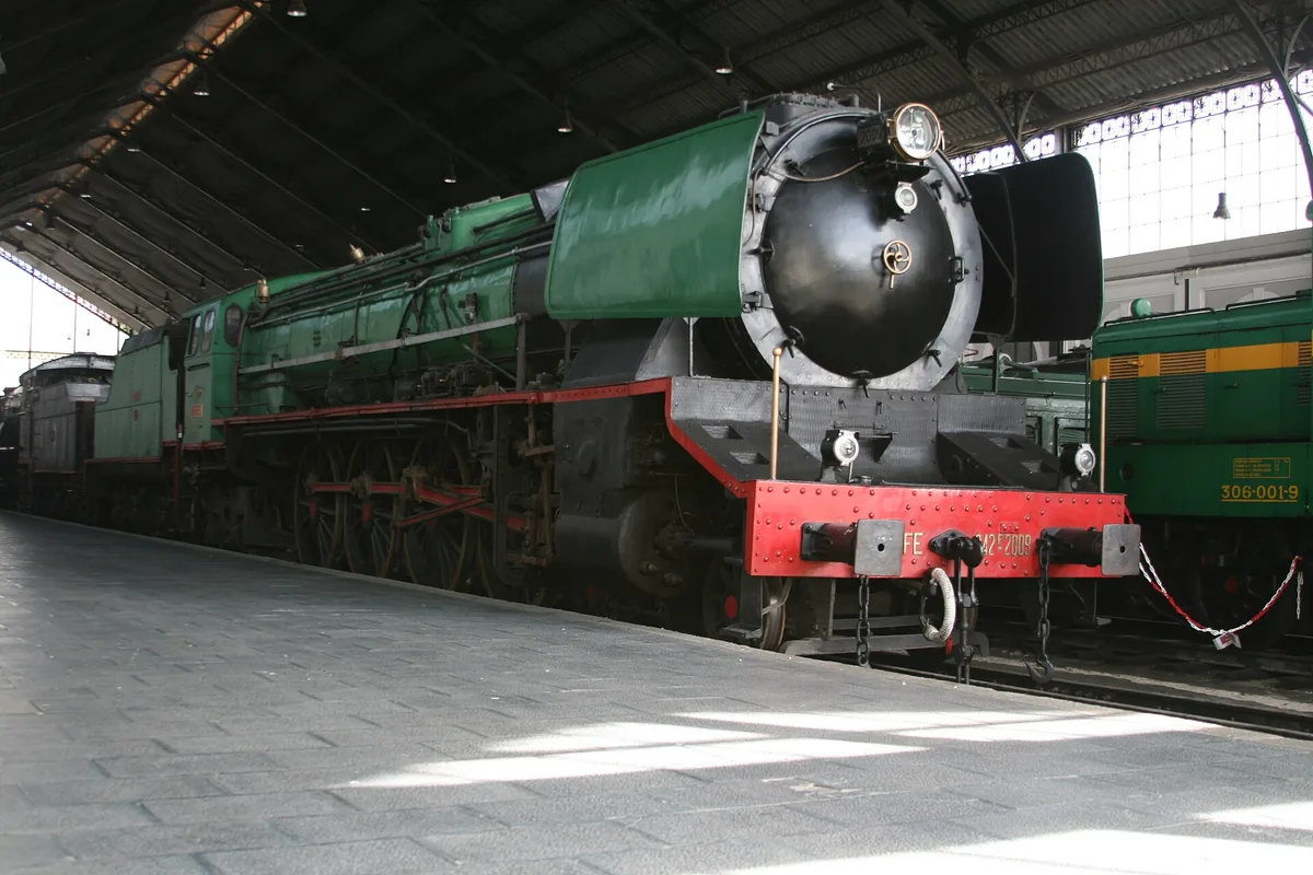 A vintage green steam locomotive on a track beneath the iron and glass roof of a railway hall