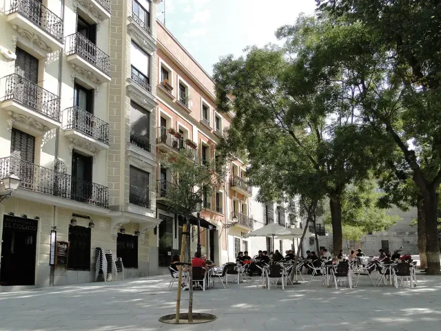 A sloping paved square with pale buildings, trees and waiting horse carriages