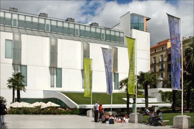 A white museum building with colourful exhibition banners at the entrance and visitors sitting on the steps