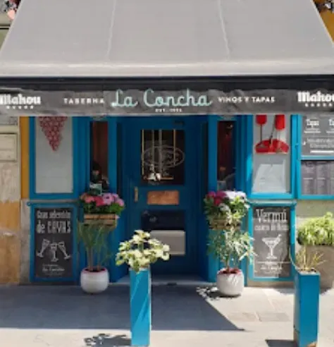A tavern facade painted light blue with an awning, potted plants at the entrance and chalkboards on both sides