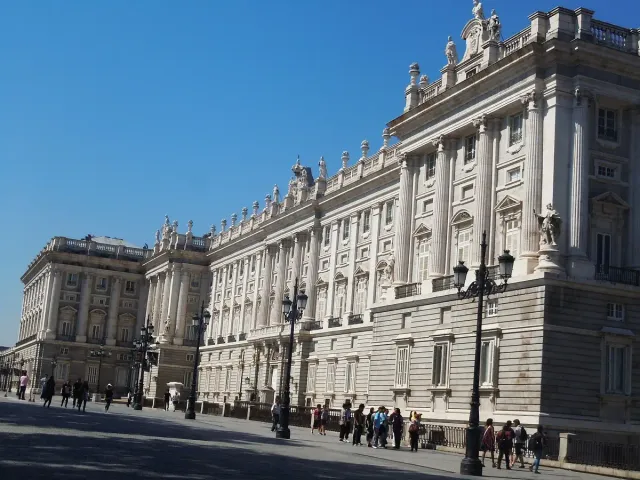 A long white stone palace with rows of columns and windows, with visitors waiting on the forecourt