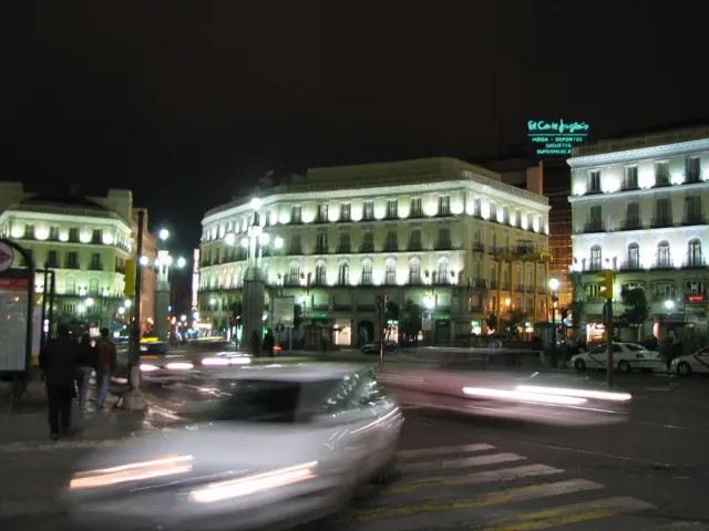 A city square at night with lit buildings and light trails from passing cars
