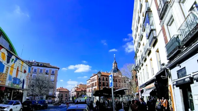 A wide street with iron balconies, stalls and the dome of a building behind under a blue sky