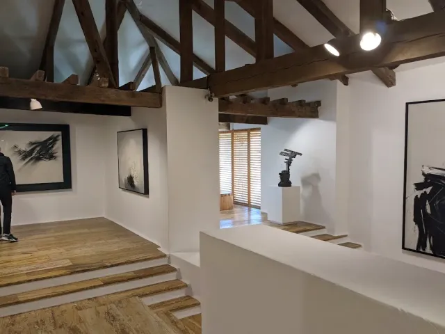 A white gallery room with dark wooden ceiling beams, abstract paintings and a small sculpture