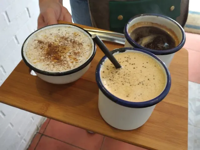 A hand holding a wooden tray with three enamel cups of coffee with foam and cinnamon