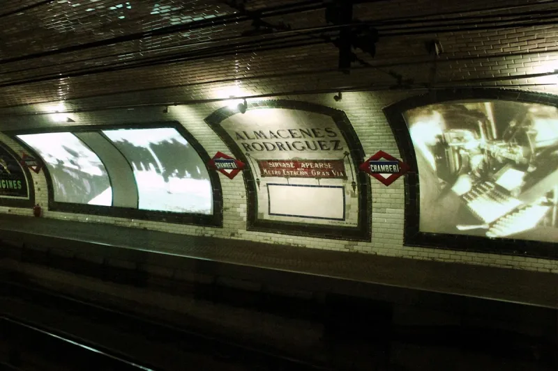 The white tiled platform wall of an abandoned metro station with vintage advertisements and two red Chamberi signs