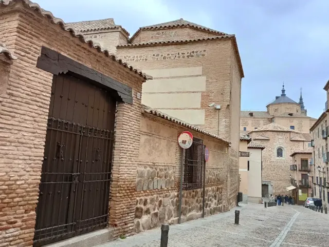 A sloping stone alley with a heavy wooden gate in a brick wall, and a dome above the rooftops behind